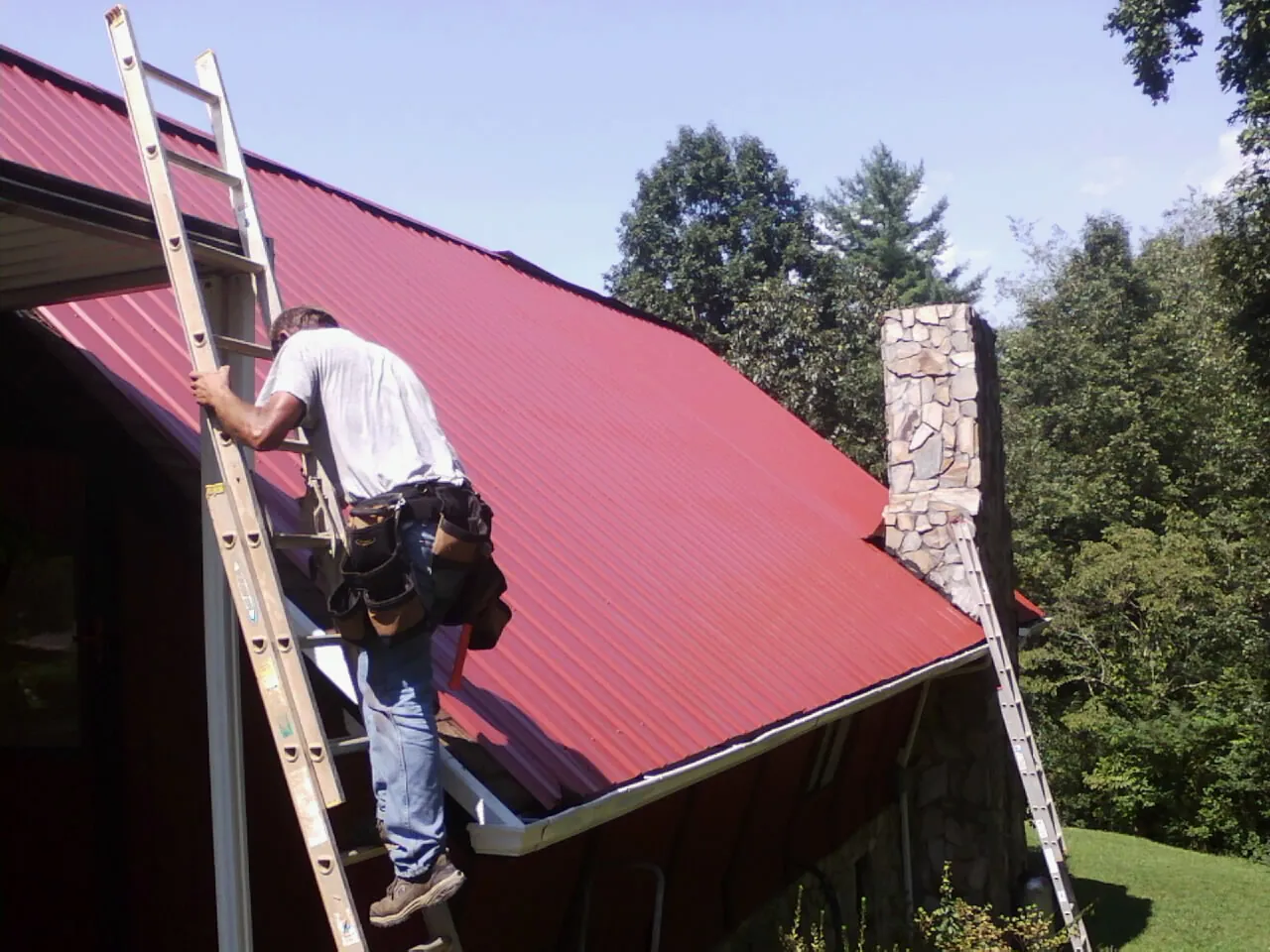 Roofer on a ladder beside a red metal roof and stone chimney — Soffit & Fascia Repair by Roofing Brothers in Eleva